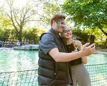A couple pose to take a selfie in front of the penguin pool holding champagne glasses