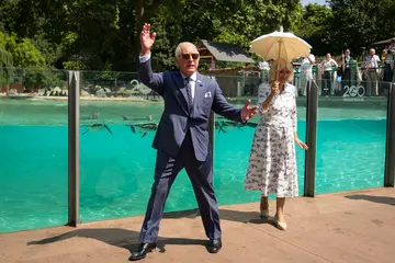 His Majesty King Charles III and Her Majesty Queen Camilla holding a parasol stand in front of glass wall at London Zoo's penguin beach.