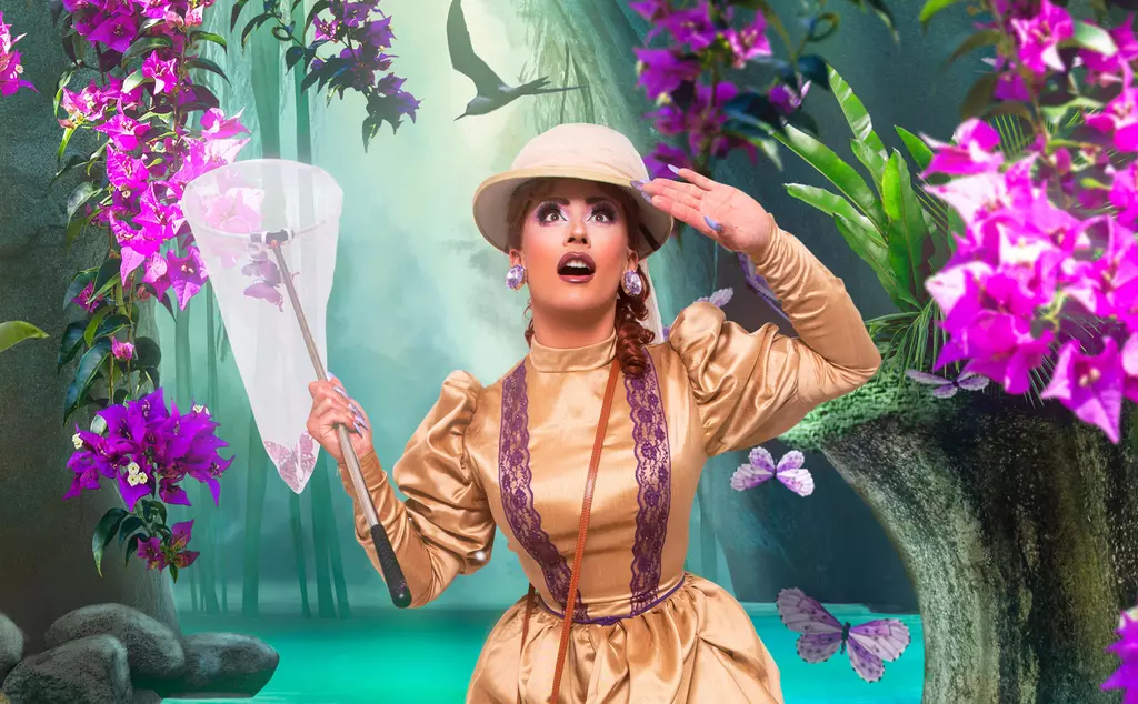 Ella Vaday, the drag queen, wears a Zoo Keeper uniform and holds a butterfly net whilst standing in front of an artificially generated lake and grass background