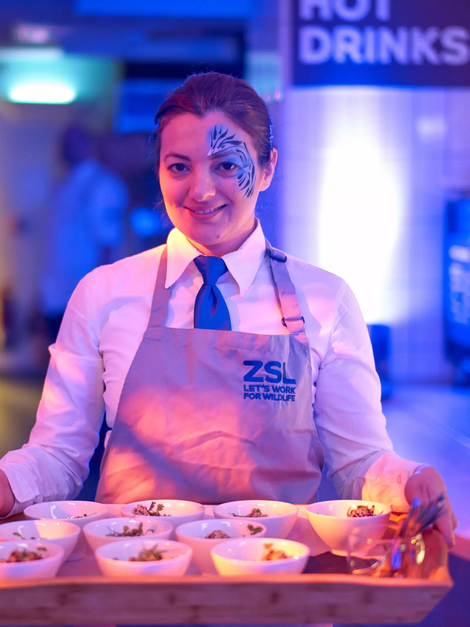 A young woman with facepaint on the left side of her face holding a tray of bowl food at an evening event