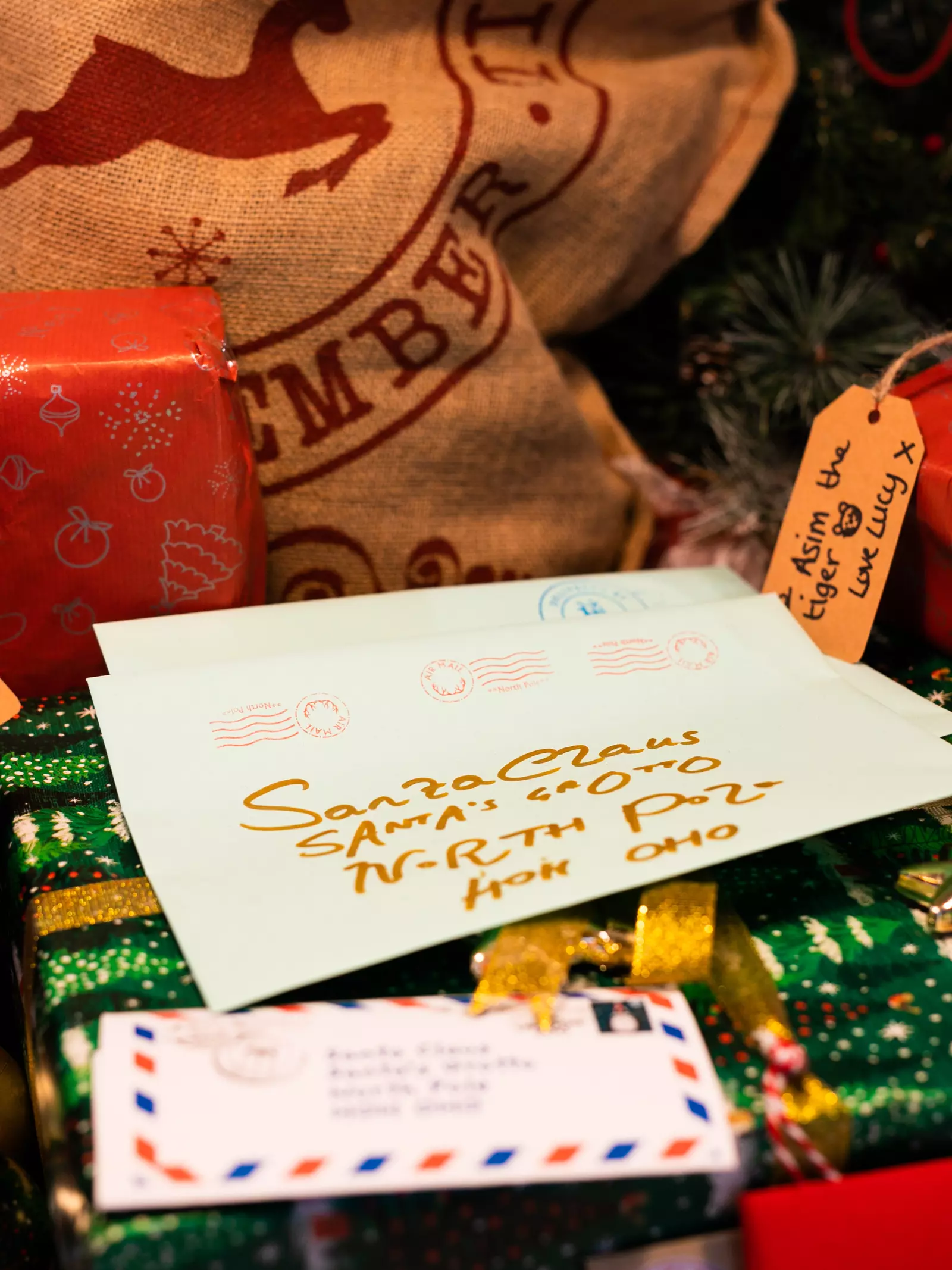 A white letter in the centre of the frame is addressed in gold lettering to Santa Claus at the North Pole. The letter is surround by red and green wrapped Christmas presents and a hessian sack