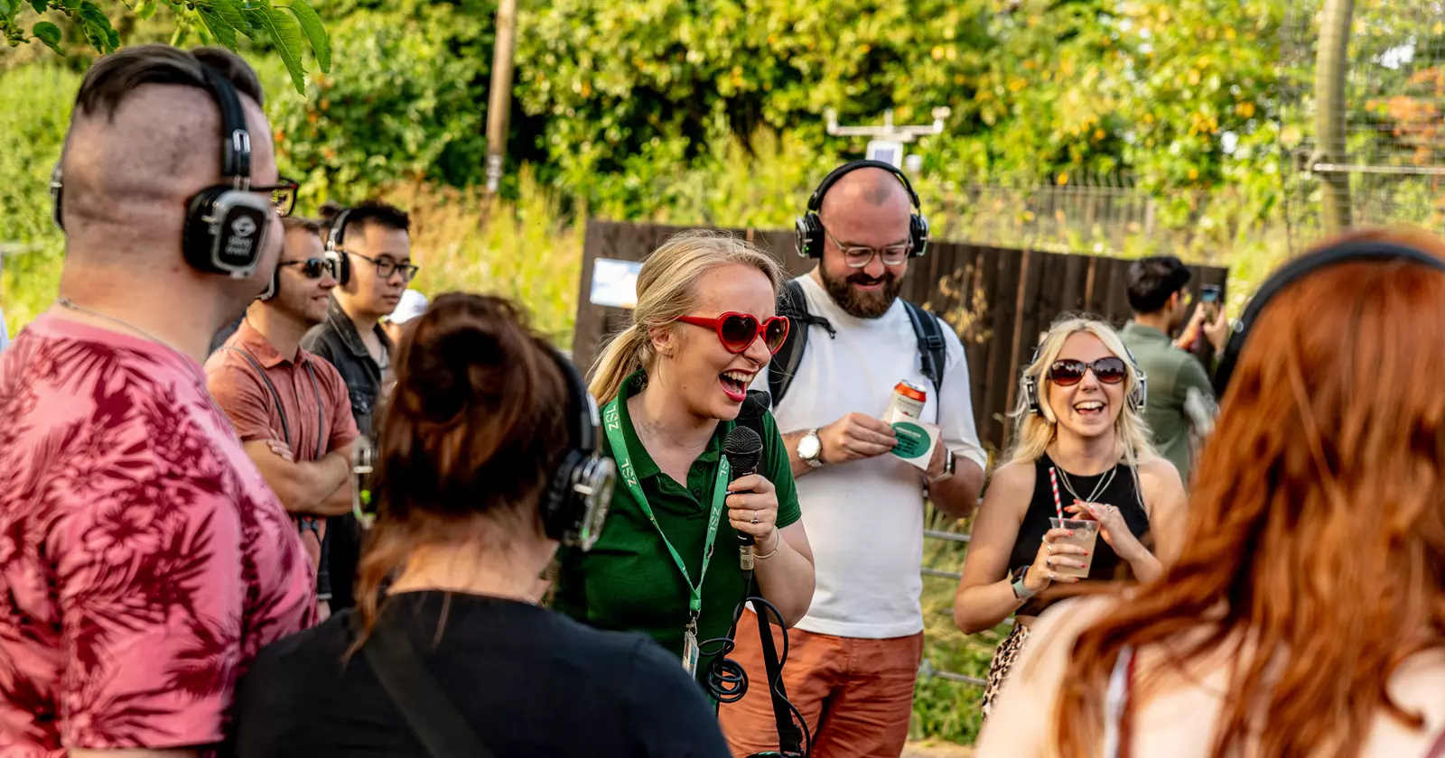 A woman in a green London Zoo t-shirt laughs into a microphone while surrounded by laughing tour guests