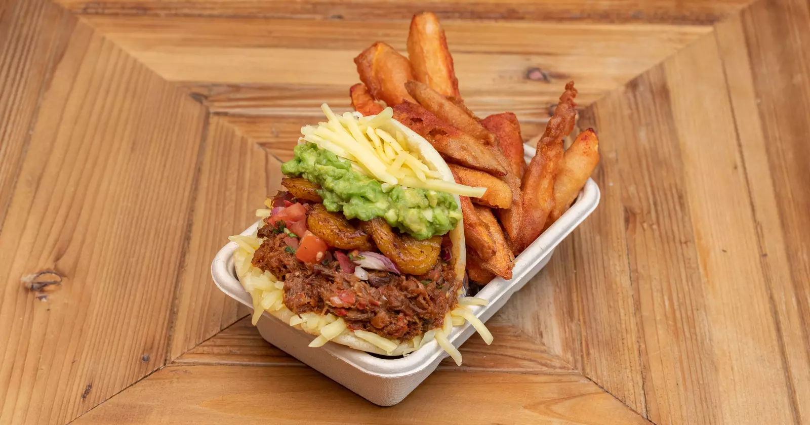 A stacked flatbread containing layers of meat, tomatoes, grilled vegetables, guacamole and cheese sits in a takeaway box next to crispy fries