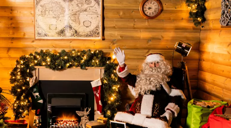 Santa sits in his warm, cosy grotto, decorated with foliage, twinkly lights and a roaring fireplace