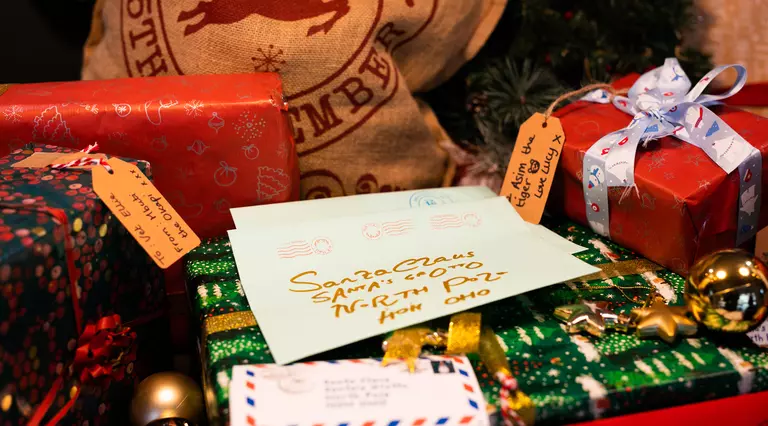 A white letter in the centre of the frame is addressed in gold lettering to Santa Claus at the North Pole. The letter is surround by red and green wrapped Christmas presents and a hessian sack