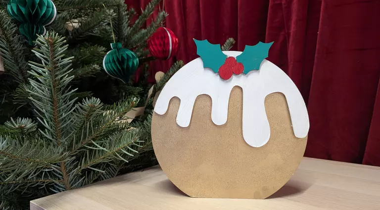 A golden Christmas pudding made of wood is framed int he background by a Christmas tree and red velvet curtain