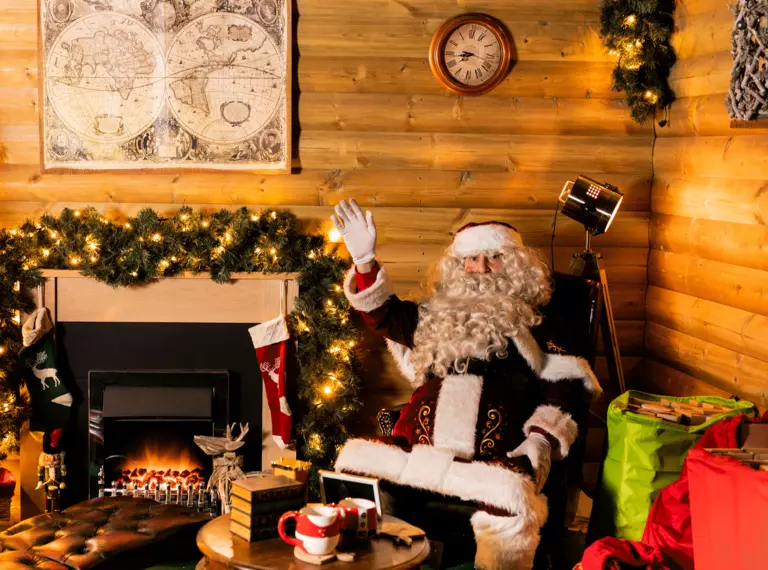 Santa sits in his warm, cosy grotto, decorated with foliage, twinkly lights and a roaring fireplace