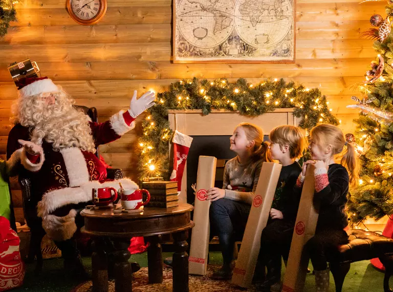 Santa sits in his warm, cosy grotto across from three young, caucasian children each holding a long rectangular present and smiling. The wooden grotto is decorated with foliage, twinkly lights and Mr and Mrs Claus mugs on the table in front of them