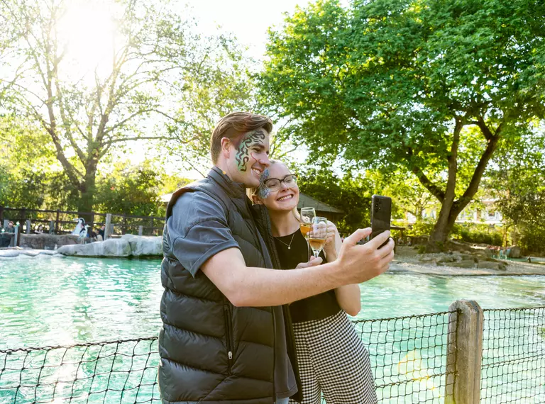 A man and a woman stand taking a selfie with their champagne glasses in front of the cool blue penguin pool at London Zoo on a summer's evening