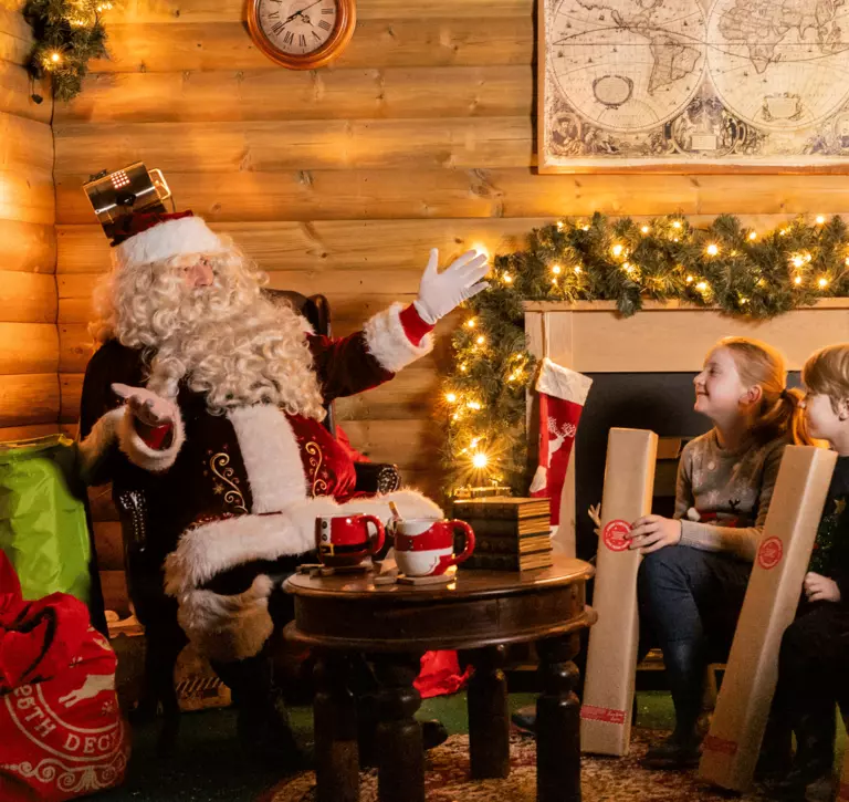 Santa sits in his warm, cosy grotto across from three young, caucasian children each holding a long rectangular present and smiling. The wooden grotto is decorated with foliage, twinkly lights and Mr and Mrs Claus mugs on the table in front of them