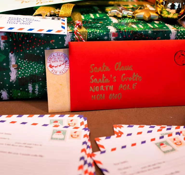 In the background of this image are piles of letters and presents addressed to Santa Claus, and in the foreground are smaller piles of blank letters that children can write to Santa